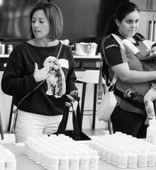 Two women standing behind a table with stacks of white containers, one wearing a sweater with a dog design.