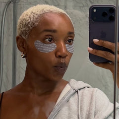 Woman with eye patches taking a mirror selfie in a bathroom.